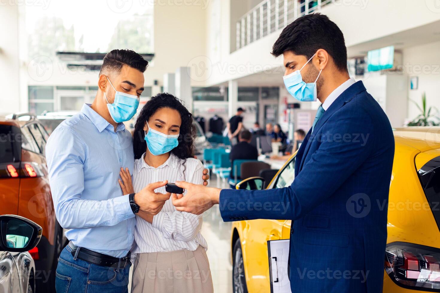 Car purchasing while coronavirus pandemic concept. Arabic couple in protective face masks buying new auto at showroom, taking key from automobile from sales manager, side view photo