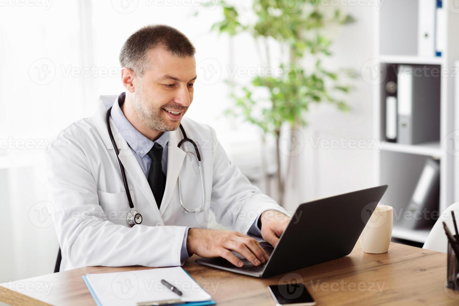 Doing Research Concept. Portrait of smiling middle aged specialist physician using computer laptop, typing on keyboard, sitting at table in hospital, giving online consulation, telemedicine photo