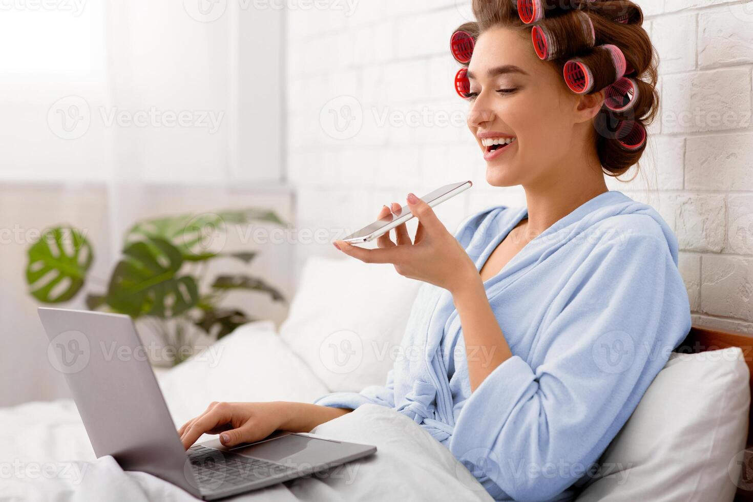 Cheerful Glamorous Woman Using Voice Search App On Cellphone And Laptop Working Online Sitting In Modern Bedroom Indoor. Mobile Technology And Communication, IoT Concept. Side View photo