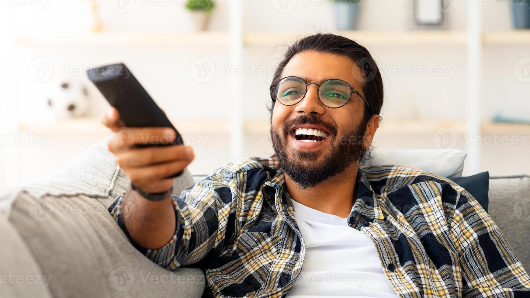 Relaxed arab guy wathcing movie on TV at home, sitting on couch alone, having happy face expression. Happy young man with TV remote watching comedy and laughing, copy space, closeup photo