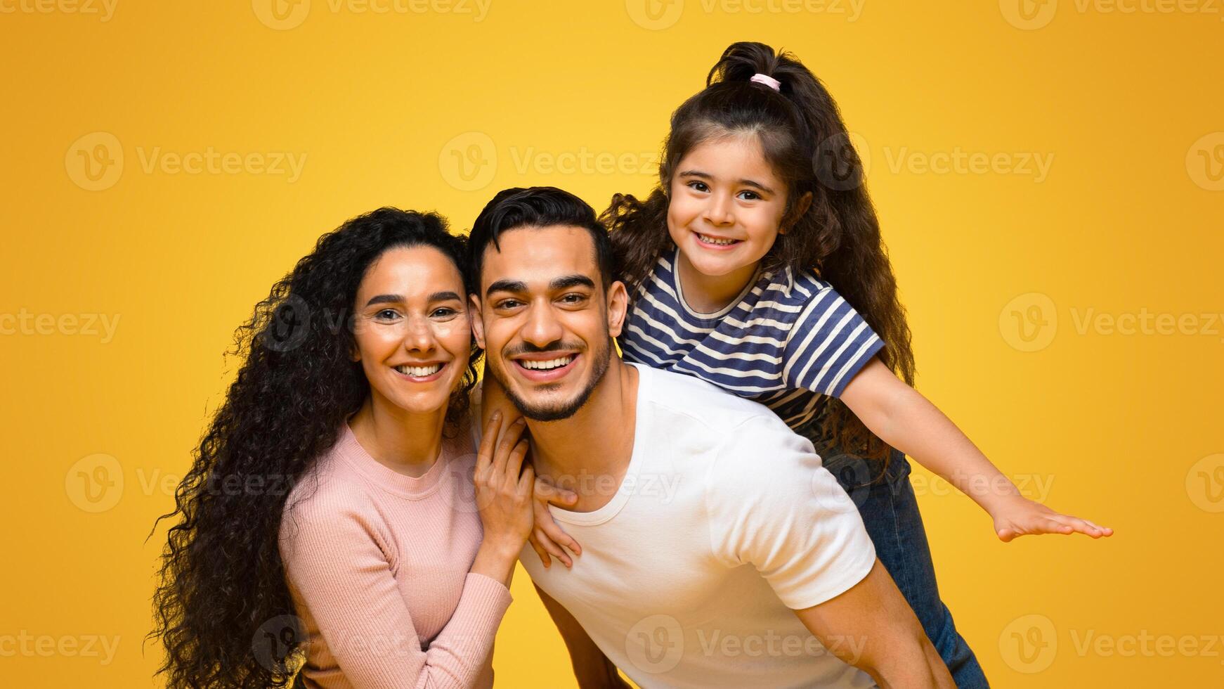 Family Time. Portrait Of Happy Arab Parents And Daughter Having Fun Together While Posing Over Yellow Background In Studio, Positive Middle Eastern Mom, Dad And Child Smiling At Camera, Copy Space photo
