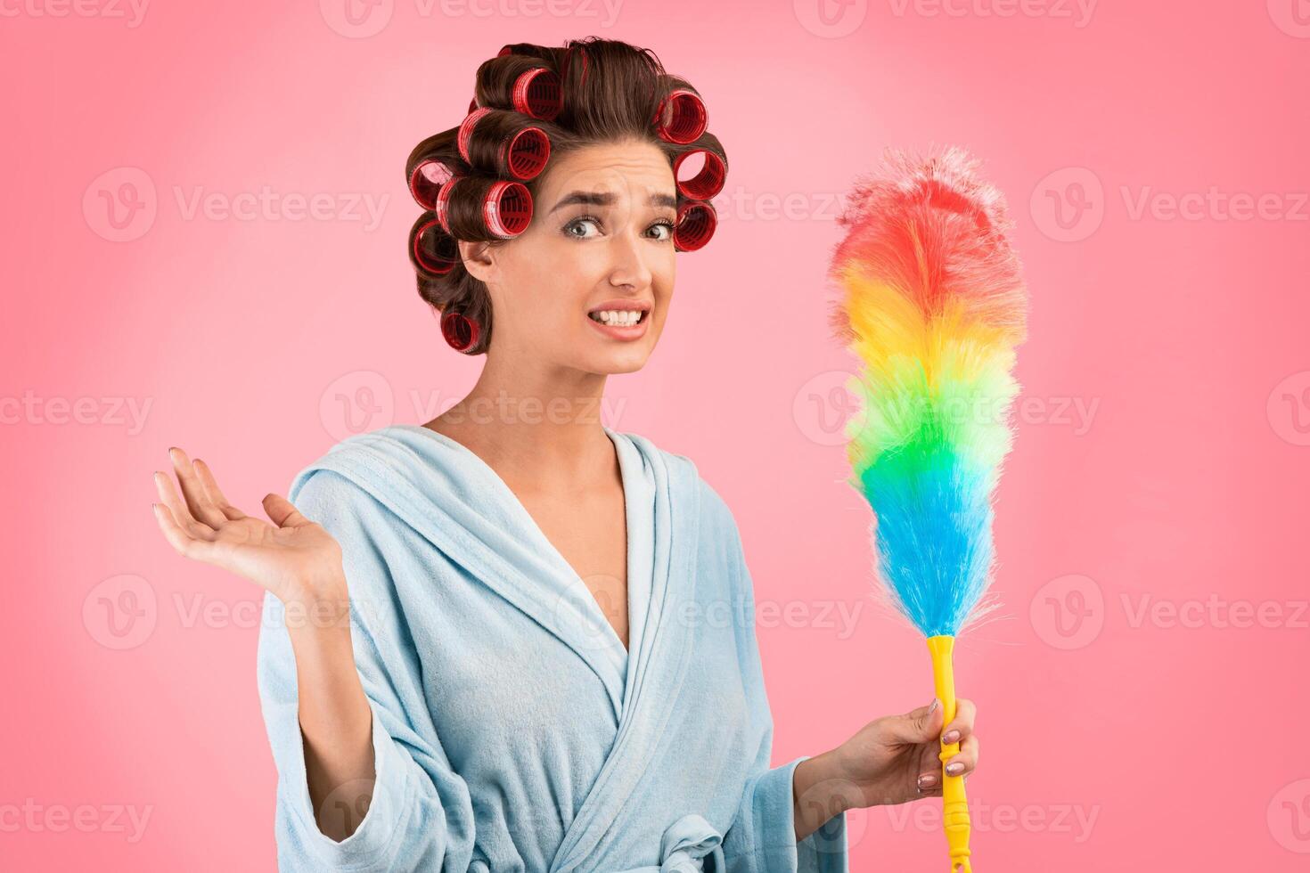 Spring Cleaning Concept. Confused Housewife With Curlers Holding Feather Duster Standing Over Yellow Background. Studio Shot photo