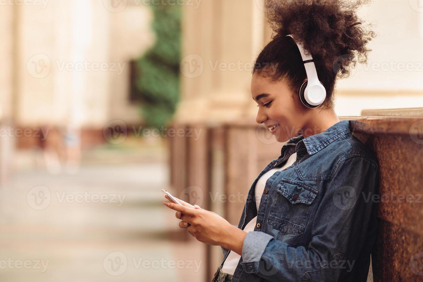 African American Female Student Girl Using Smartphone Texting Wearing Earphones And Browsing Internet At College Outdoors. Educational Mobile Application, E-Learning Concept. Side View Shot photo