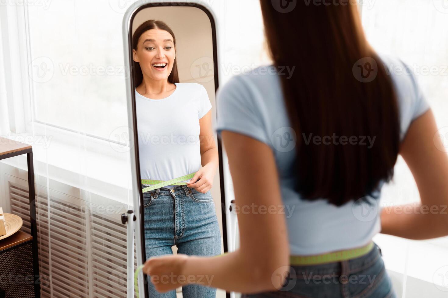 Weight Loss. Happy Girl Measuring Waist With Tape Standing In Front Of Mirror Slimming At Home. Copy Space, Selective Focus photo