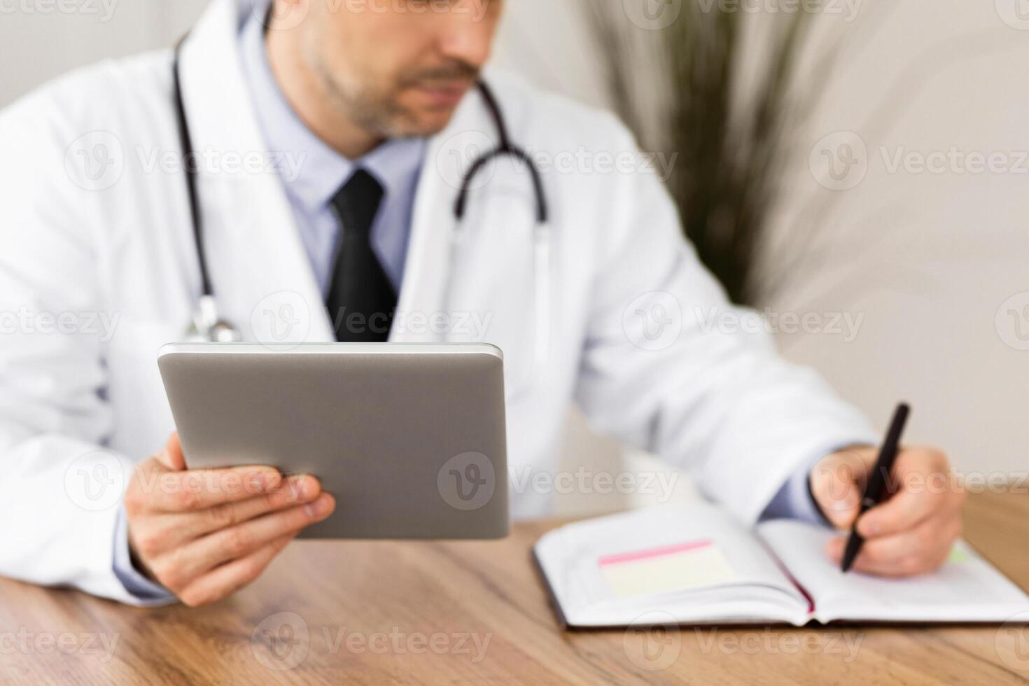 Healthcare, Medical Care And Technology Concept. Male doctor working in office at clinic, writing with pen in notebook, holding and using digital tablet, checking appointment schedule photo
