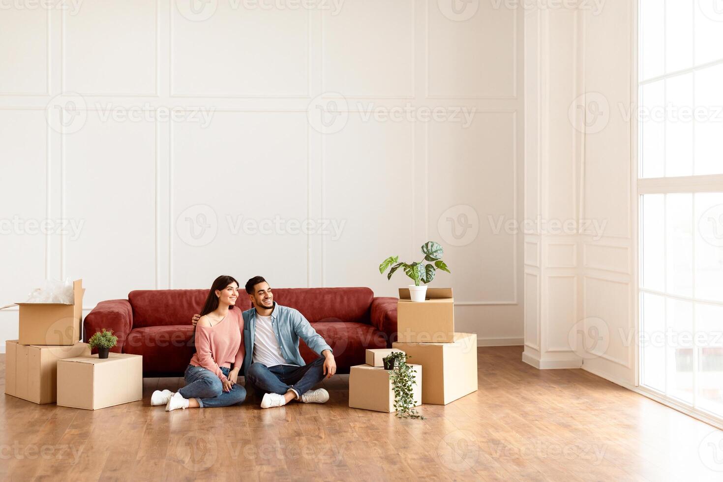 Moving House. Cheerful Middle Eastern Spouses Sitting Among Unpacked Moving Boxes In New Home, Hugging And Looking Aside At Window. Real Estate Ownership And Apartment Rent Concept. Free Copy Space photo