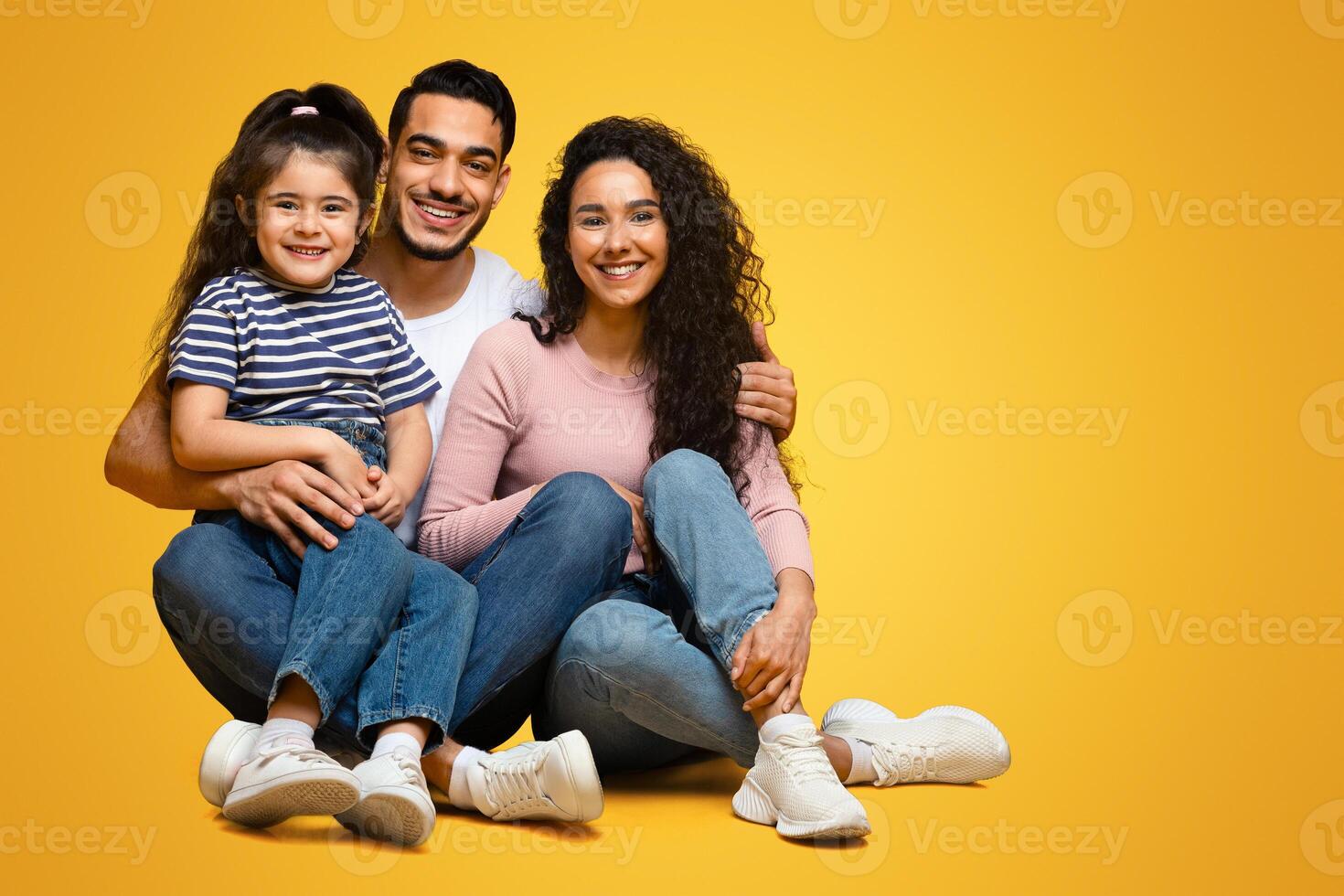 Happy middle eastern family sitting on floor and embracing, cheerful loving arab parents posing together with their cute little daughter over yellow studio background, looking at camera, copy space photo