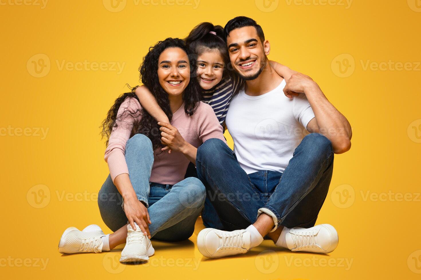 Portrait Of Young Happy Middle Eastern Family With Little Daughter Posing Together, Cheerful Arab Parents And Their Child Embracing While Sitting On Floor Over Yellow Background In Studio, Free Space photo