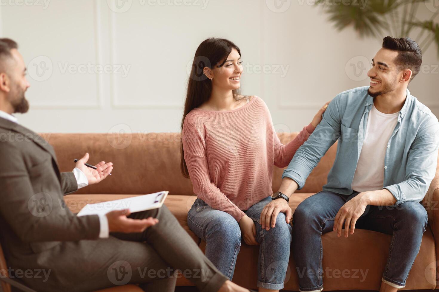 Couples Therapy Concept. Happy husband and wife reconciling at session in therapists office during appointment, looking at each other, smiling woman touching man's shoulder, sitting on couch sofa photo