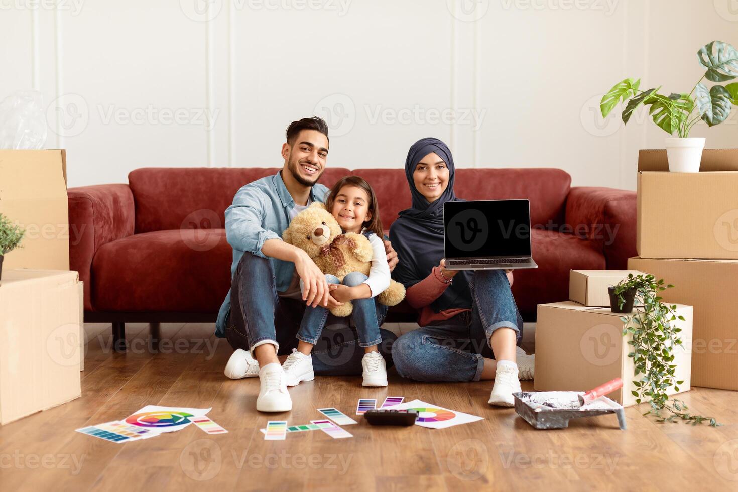 Great Website. Smiling muslim dad, mom and daughter sitting on floor near sofa in living room among carton boxes and paint for repair, arab woman showing computer with black empty screen for mock up photo