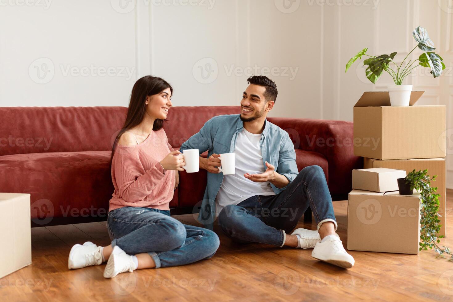 Loving husband and wife sitting on the floor leaning on couch in living room, having break while unpacking stuff at new house, talking and drinking coffee. Moving, recolating concept photo