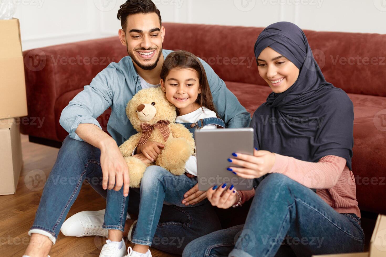 Relocation Concept. Joyful Middle Eastern Family Of Three People Using Tablet Computer Sitting On Floor Among Moving Boxes In New House, Choosing And Ordering Furniture, Discussing Decor photo