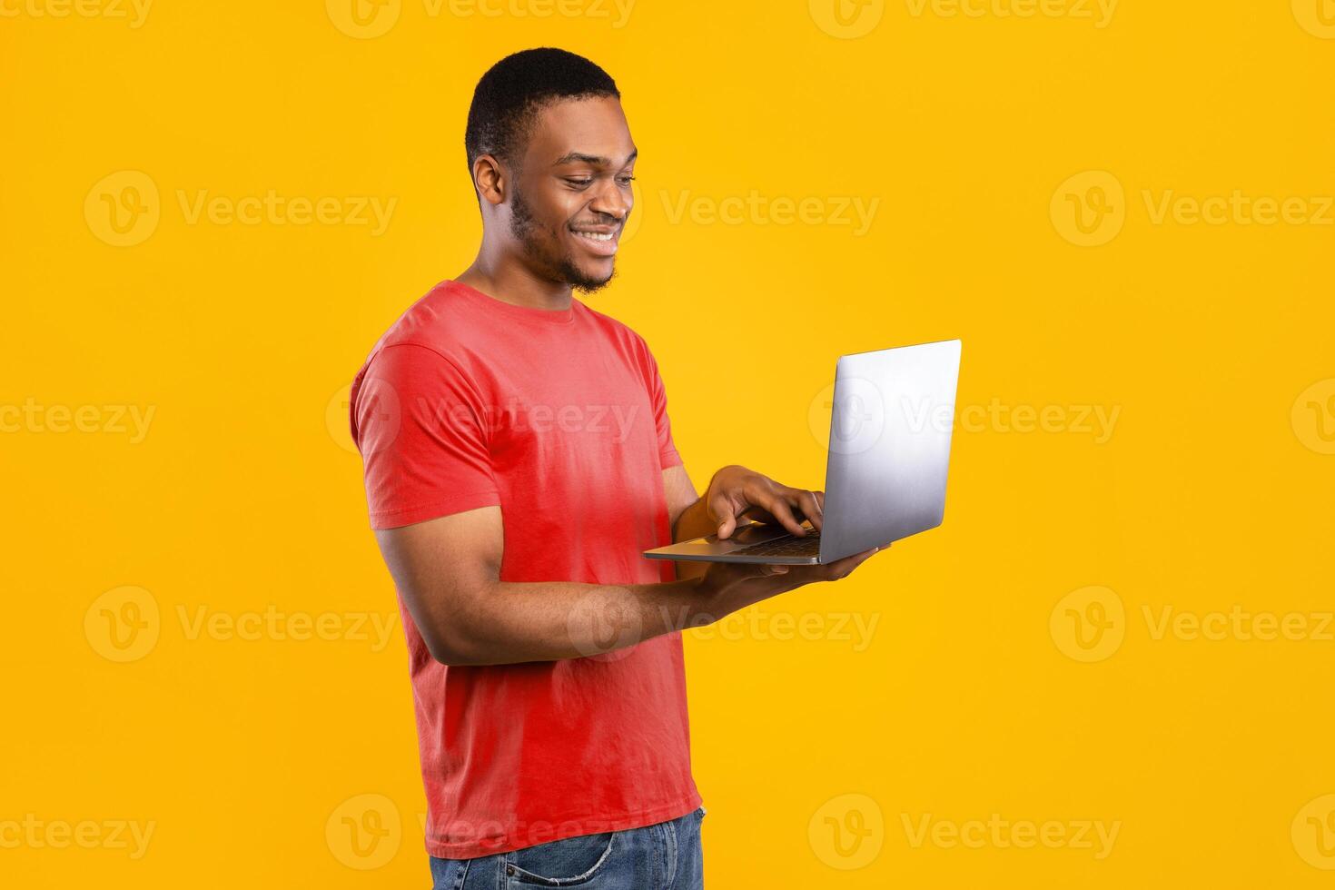 Happy African American Freelancer Man Using Laptop Standing In Studio Over Yellow Background. Black Guy Browsing Internet Holding Computer. E-Learning, Technologies Concept. Side View photo