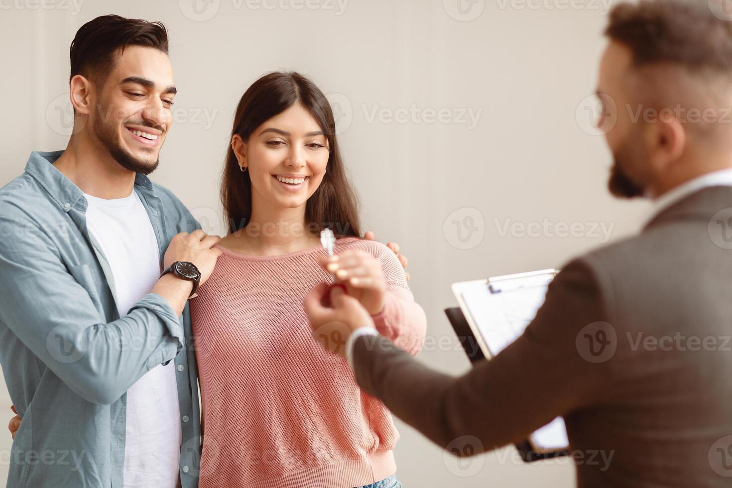 Property For Sale Concept. Portrait of happy smiling couple taking key of future new apartment. Professional real estate agent wearing suit holding in hand and giving keys to young buyers. Mortgage photo