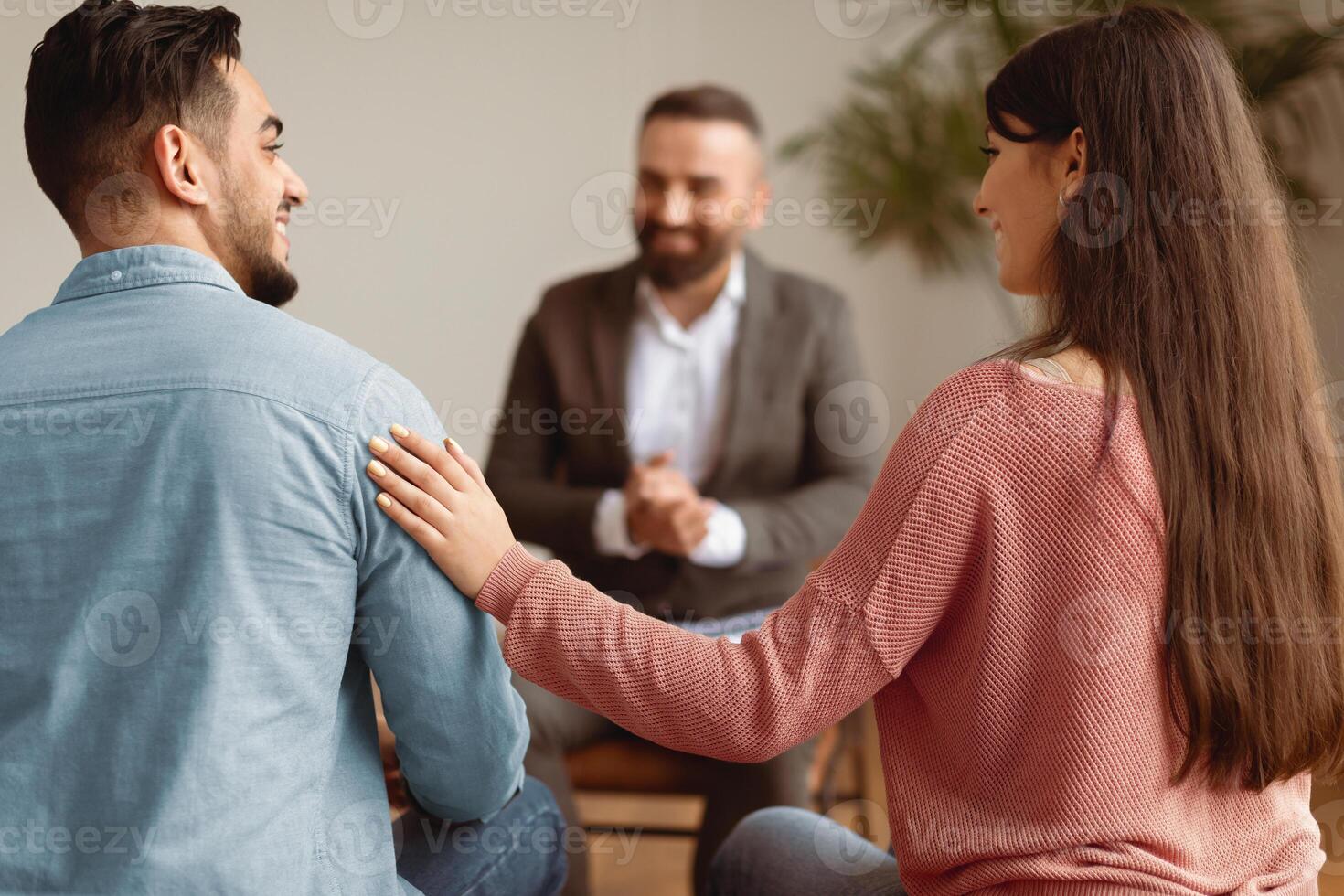 Marital Therapy Concept. Happy couple reconciling at session in therapists office during appointment, looking at each other, smiling woman touching man's shoulder, back view, selective focus photo