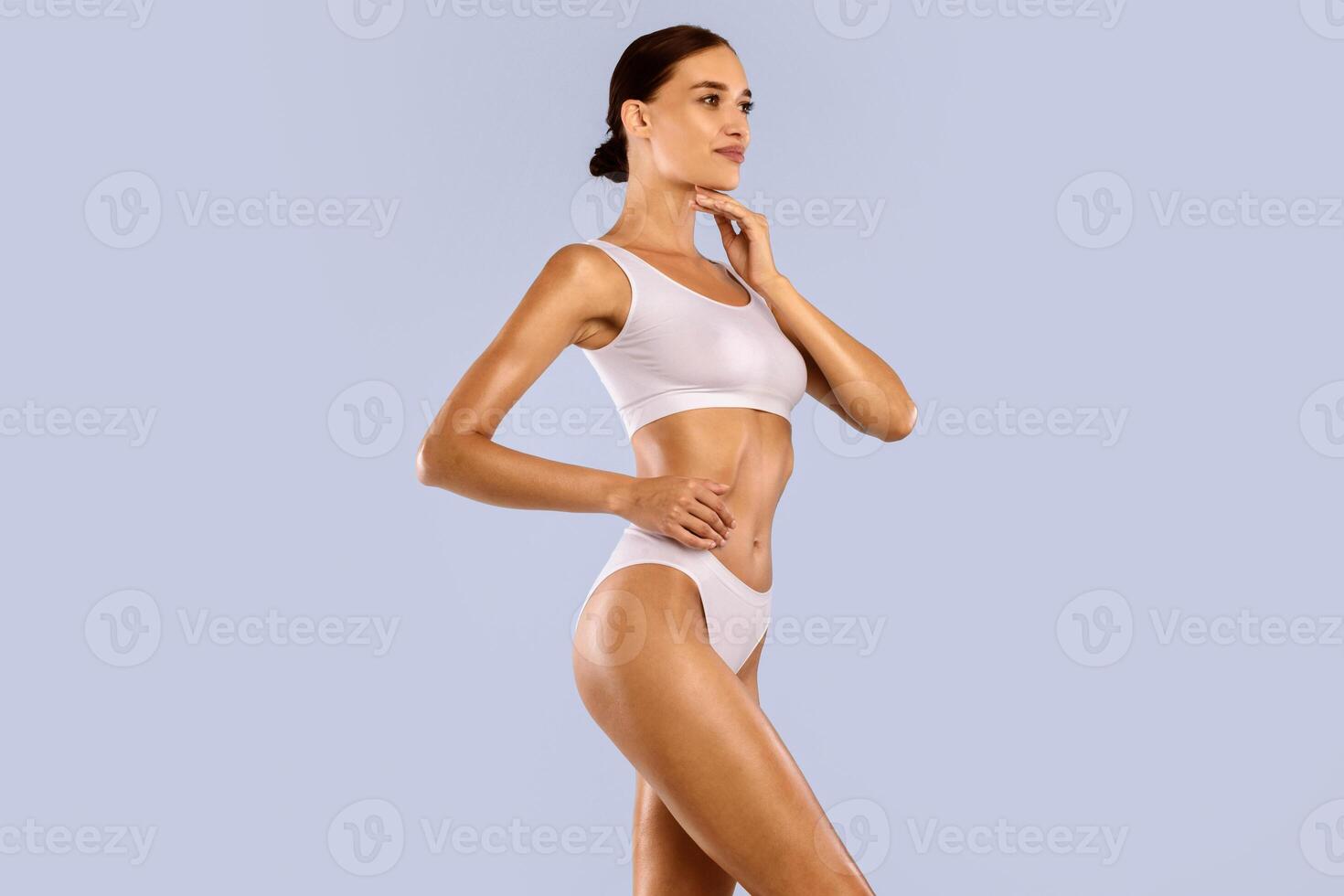 Fitness, Diet, Skincare Concept. Portrait Of Half-turned Smiling Tender Slim Lady With Perfect Ideal Body Shape Touching Soft Skin On Her Neck With Hand Posing Isolated On Blue Studio Background photo