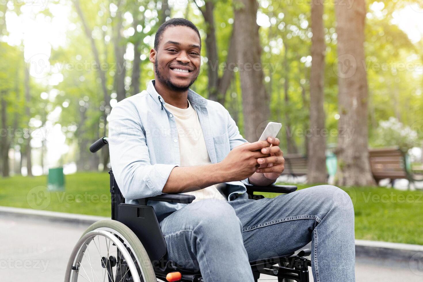 Happy disabled black man in wheelchair using smartphone, checking messages online at park. Young African American handicapped guy with mobile phone posting in social network, browsing web outdoors photo