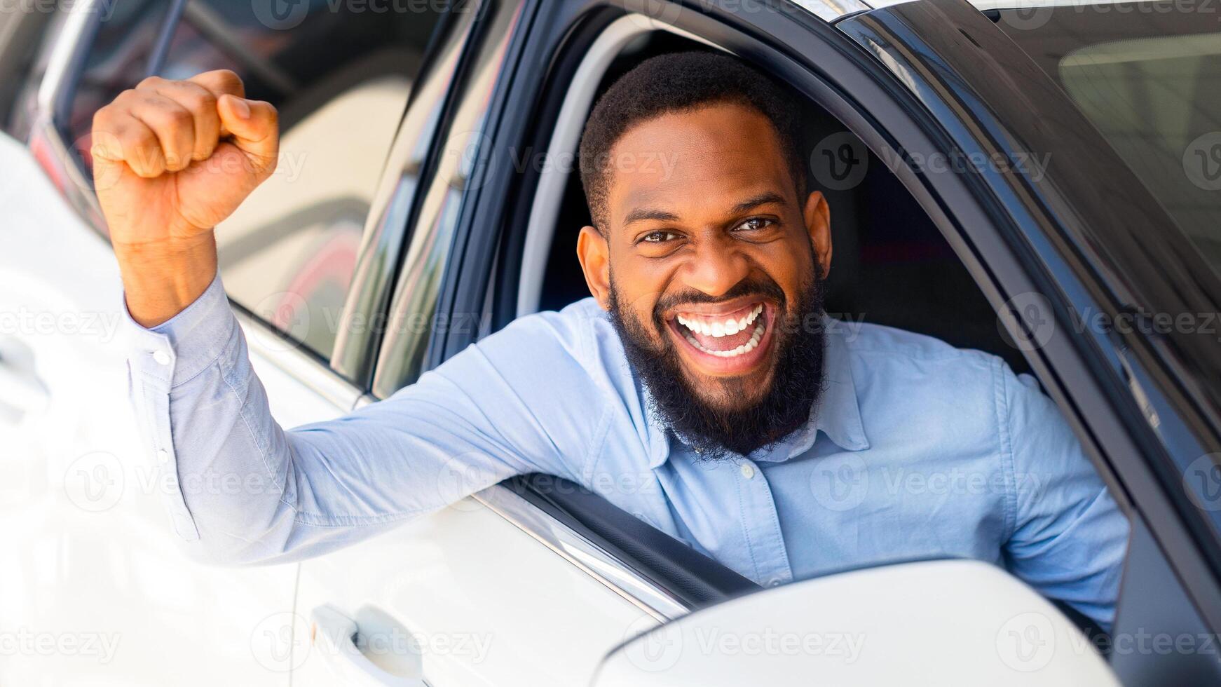 Excited Black Guy Posing In His New Car After Buying, Raising Fists And Exclaiming With Joy Through Window, Joyful African American Man Celebrating Purchasing Modern Vehicle In Dealership Center photo