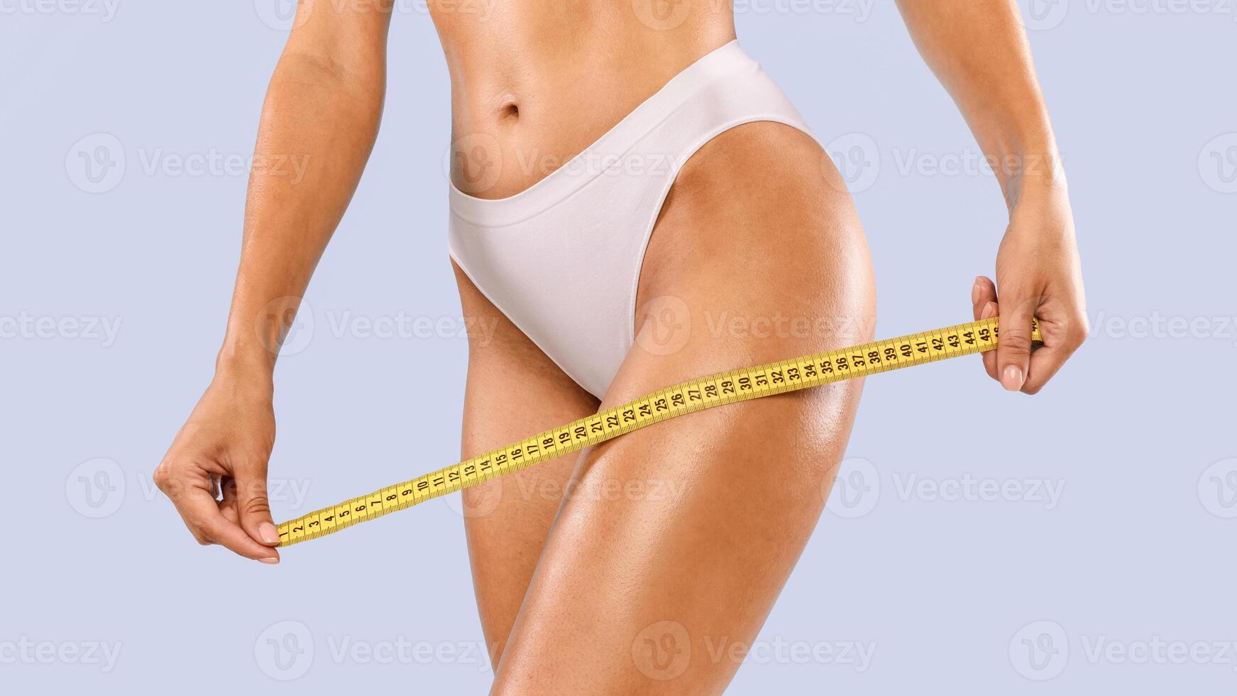 Perfect Body Concept. Cropped closeup view of slim woman in white panties holding yellow tape ruler hear hips, measuring thigh. Unrecognizable lady checking her size isolated on blue studio background photo