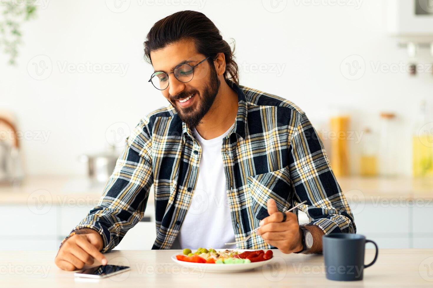 Handsome young indian long-haired, bearded man using cellphone while having healthy and tasty breakfast, checking social media or watching funny videos online, sitting at kitchen alone, copy space photo