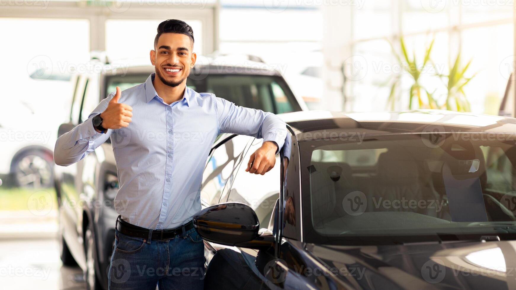 Happy arab guy standing by nice sports car in auto salon, showing thumb up and smiling at camera, rich middle-eastern young man buying new automobile in luxury showroom, copy space photo