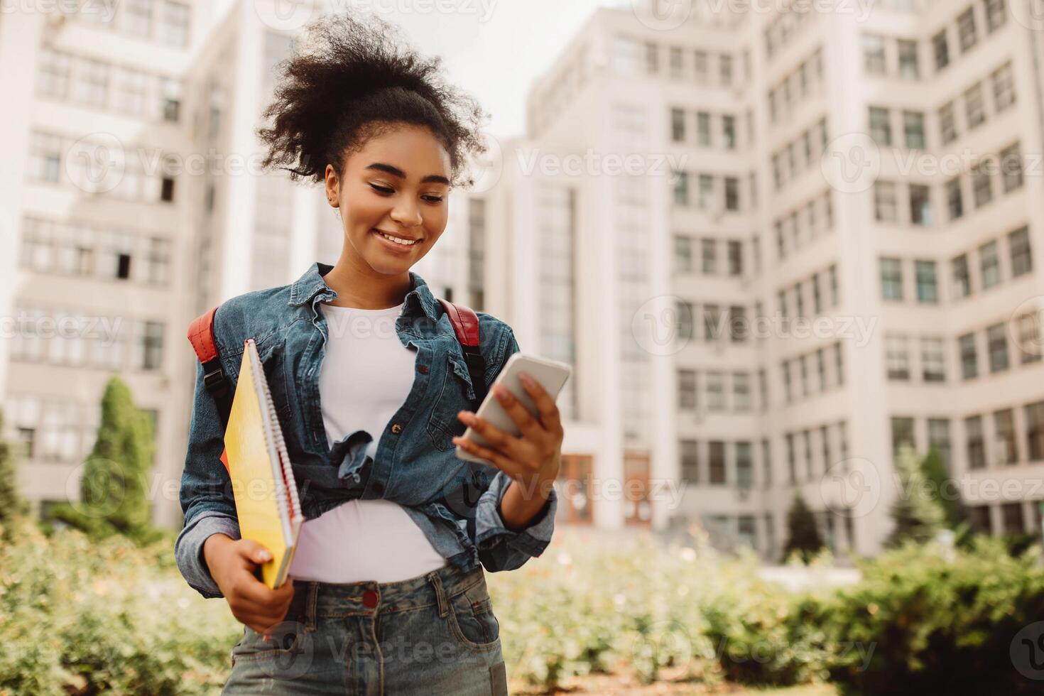 Happy African American Student Girl Using Phone With Educational App Standing Outdoors. Millennial Female Texting And Browsing Internet Via Smartphone Posing Near College Building photo