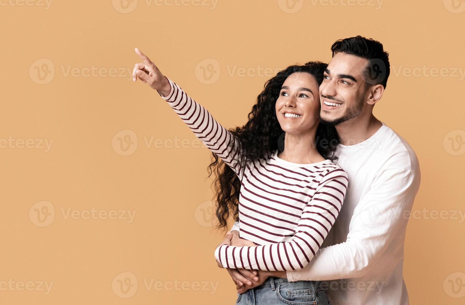 Cheerful Young Arab Couple Pointing With Finger Aside At Copy Space, Excited Millennial Middle Eastern Man And Woman Embracing And Looking At Free Place Over Yellow Background, Panorama photo