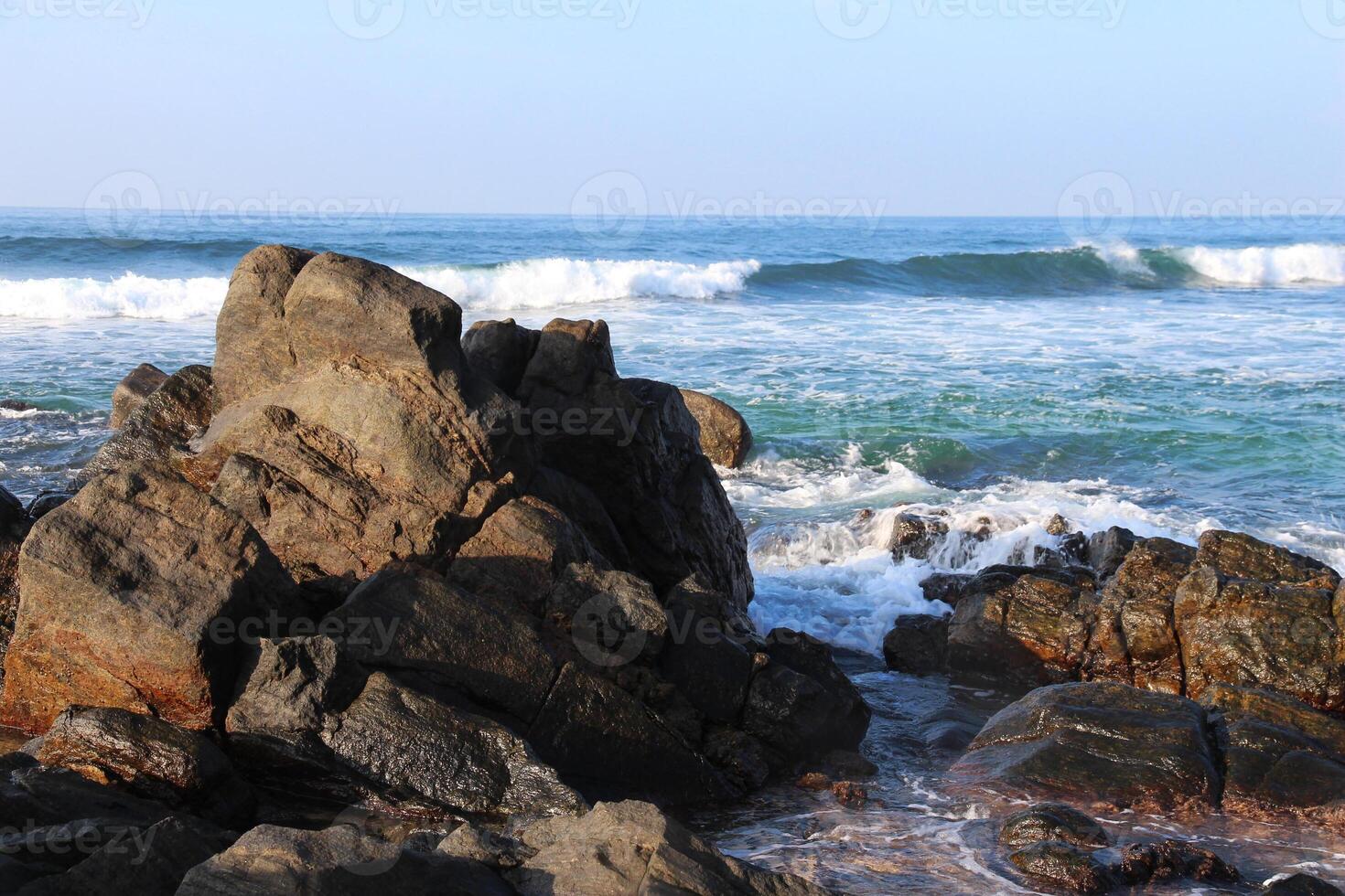 Rocky shore with waves crashing against the stones, under a clear blue sky. A scenic coastal landscape showcasing the beauty of nature and the dynamic power of the ocean photo