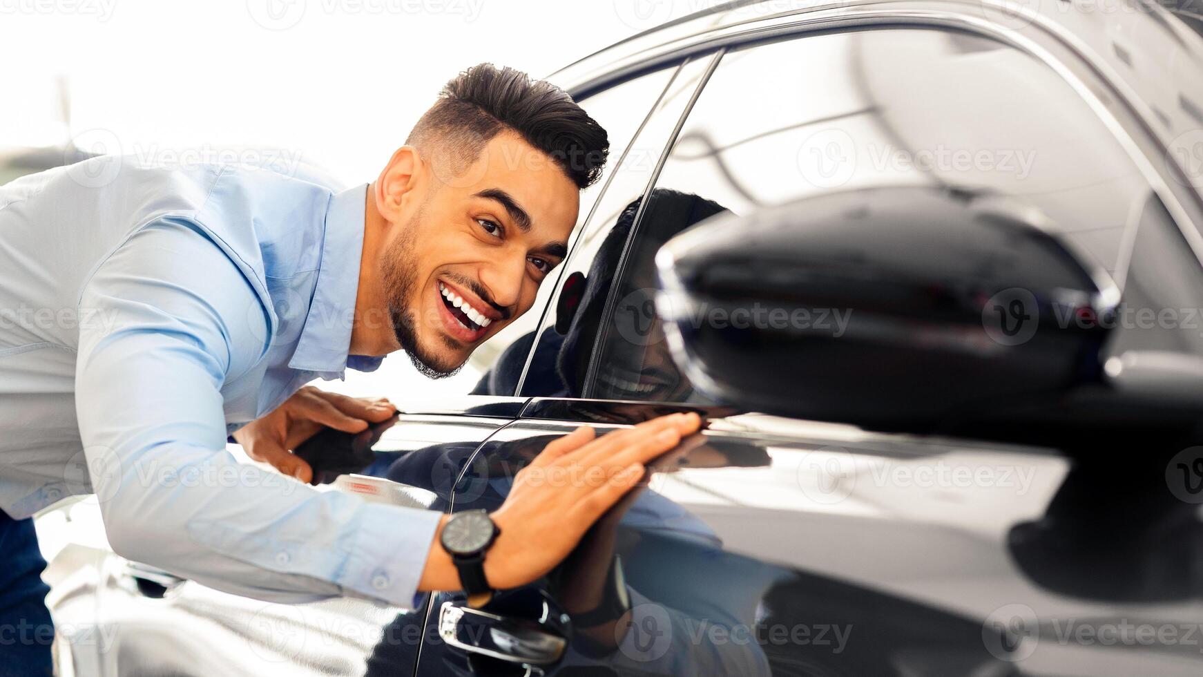 Handsome young arab man buying new car, standing at car salon and checking automobile surface, closeup. Smiling middle-eastern guy looking at beautiful black auto in showroom photo