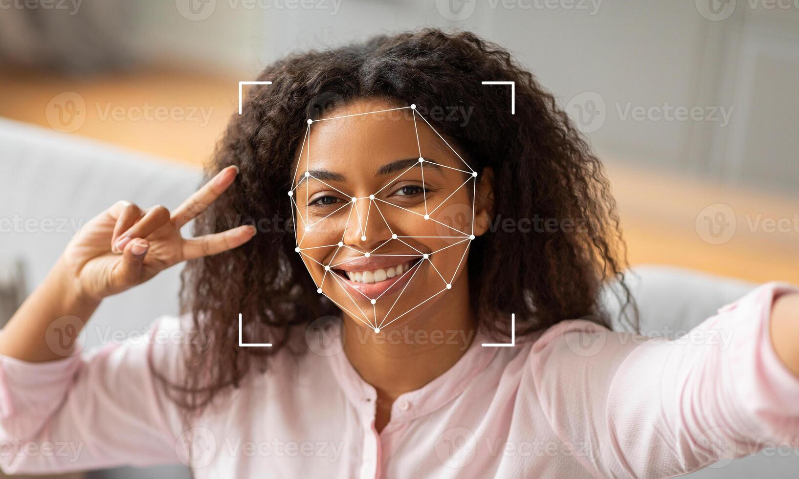 A woman with curly hair smiles and makes a peace sign, while facial identification technology analyzes her face. The indoor setting features a cozy atmosphere with soft lighting. photo