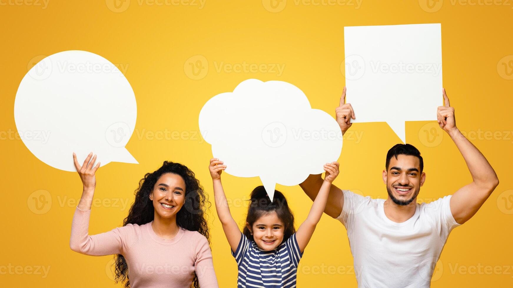 Diverse Opinions. Arab Mom, Dad And Little Daughter Holding Different Empty Speech Bubbles Above Their Heads, Middle Eastern Family Of Three Sitting On Floor On Yellow Studio Background, Copy Space photo