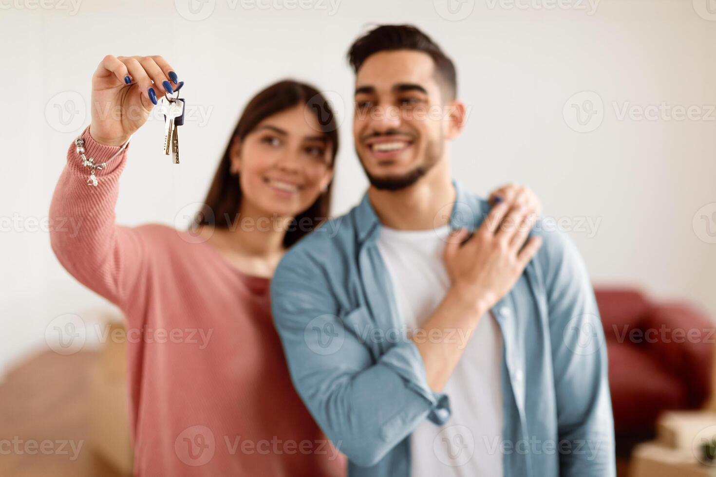 Positive young couple showing house key, standing in living room of new home, selective focus hand. Millennial arab guy and lady happy to move to bigger apartment, holding flat key, blurred background photo