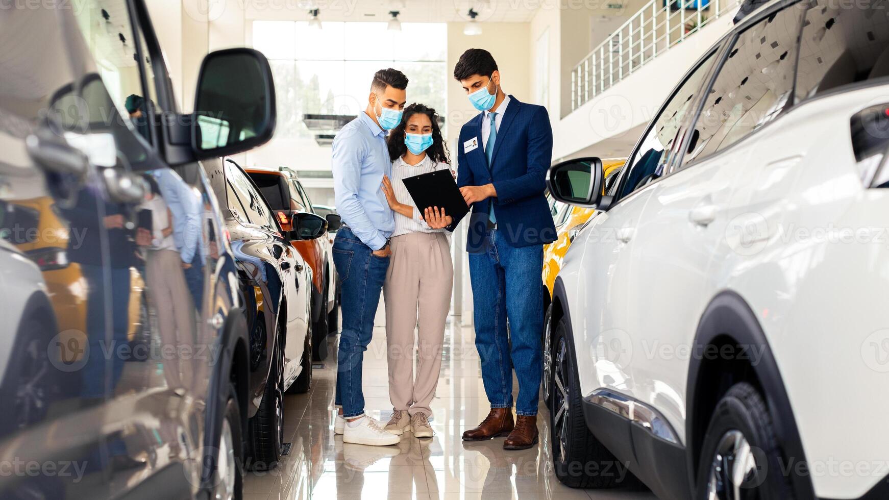 Middle-eastern young man and woman in protective face masks choosing auto in dealership salon while COVID-19 pandemic, having conversation with sales manager, reading car specifications, full length photo