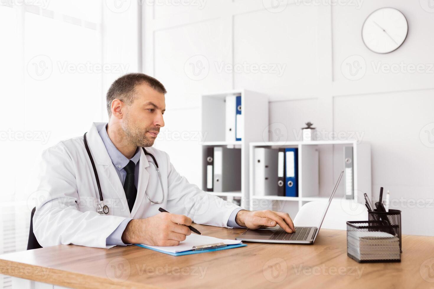 Healthcare And Telemedicine Concept. Portrait of middle aged general practitioner using computer laptop, typing on keyboard, writing with pen, doing paperwork in his office at clinic photo