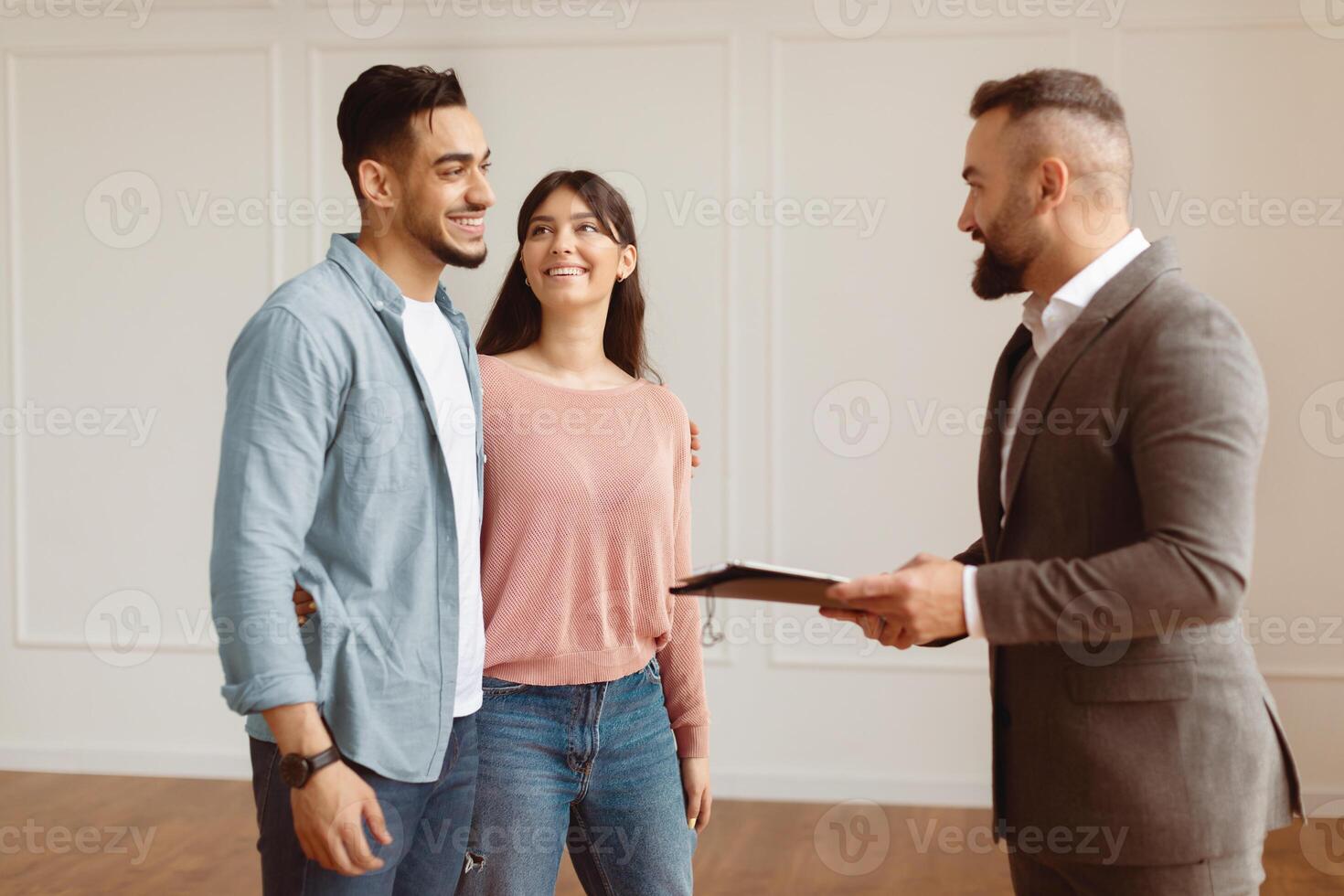 Portrait Of Smiling Professional Man Realtor In Suit Showing New Apartment To Young Arab Couple, Holding Clipboard. Happy Middle Eastern Guy And Lady Hugging And Talking With Seller photo