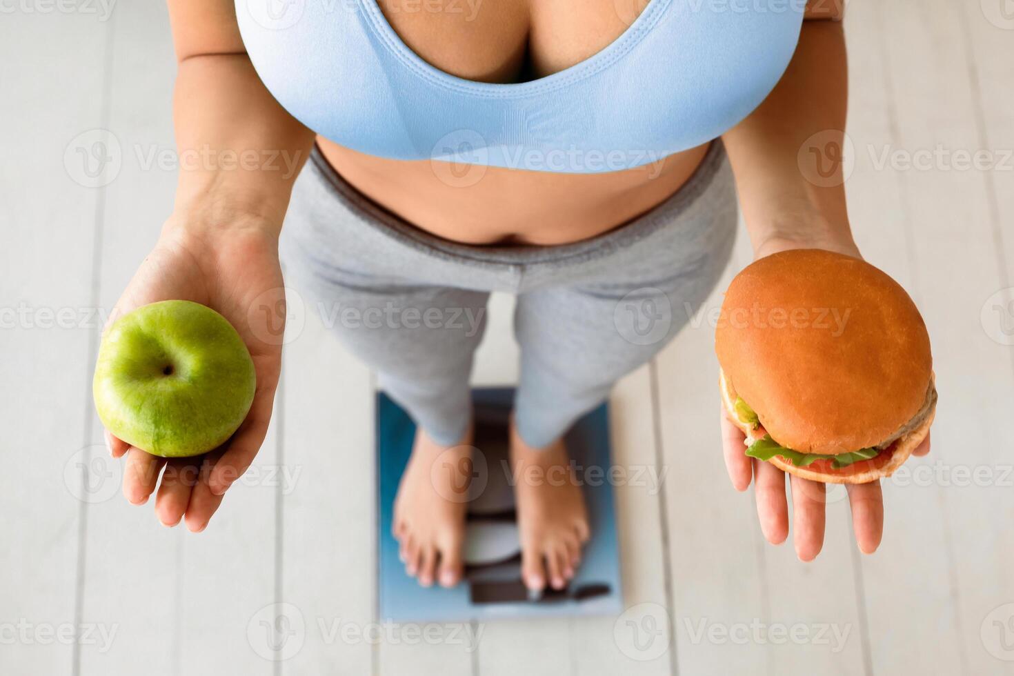 Dieting And Weight Loss. Slimming Woman Choosing Between Hamburger And Apple Standing On Weight-Scales At Home. Cropped, High-Angle photo