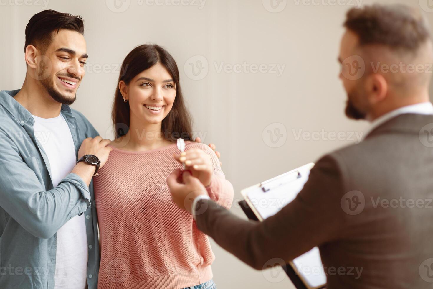 Happy smiling Middle Eastern couple taking key of future new apartment. Professional real estate agent wearing suit holding in hand and giving keys to young buyers. Mortgage, Property For Sale Concept photo
