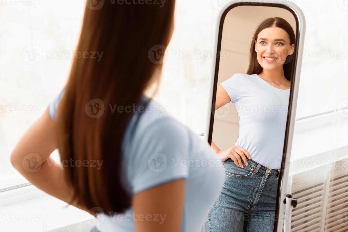 Smiling Girl Looking At Mirror After Slimming Happy About Dieting Result Standing Indoors. Weight Loss. Selective Focus photo