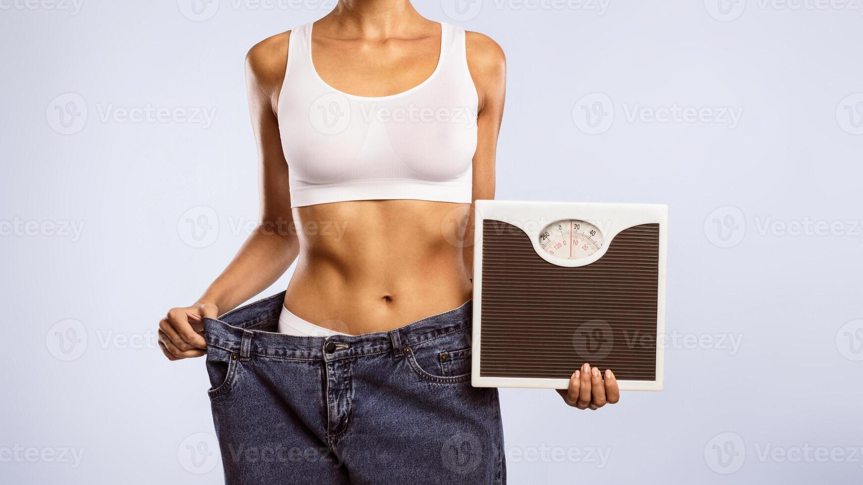 Closeup cropped view of unrecognizable woman in oversized jeans holding scales, showing results of weight loss program or liposuction. Lady achieving success in slimming, blue studio, banner, panorama photo