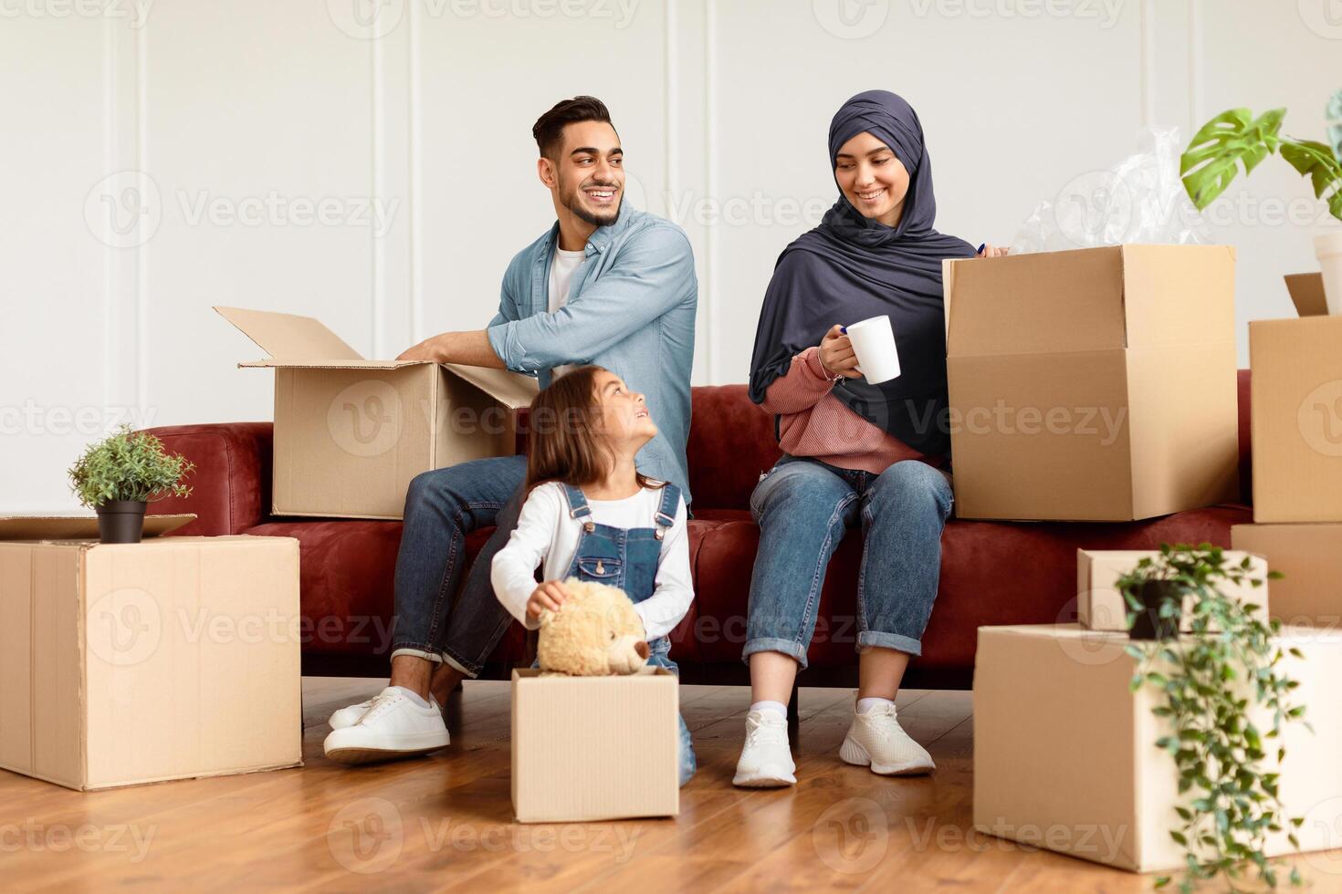 House Move And Relocation Concept. Portrait Of Cheerful Family Housing, Parent And Happy Smiling Little Girl Packing Or Unpacking Cardboard Boxes Together In Livin Room, Sitting With Open Packages photo