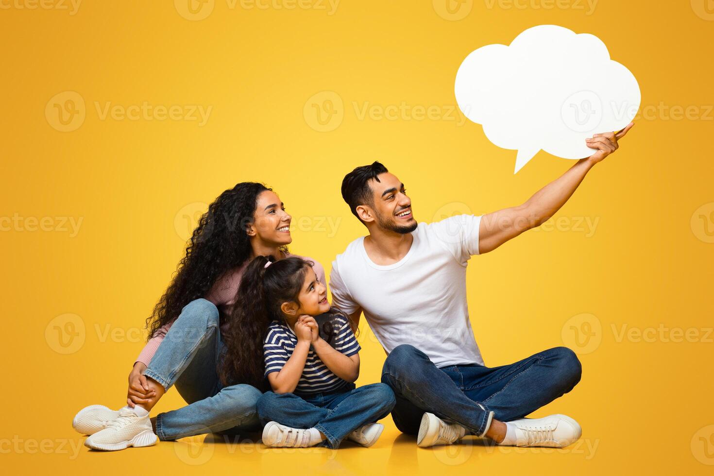 Cheerful Middle Eastern Family Of three Holding And Looking At Empty Speech Bubble While Sitting Together Over Yellow Background In Studio, Arab Parents And Child Excited About Interesting Offer photo