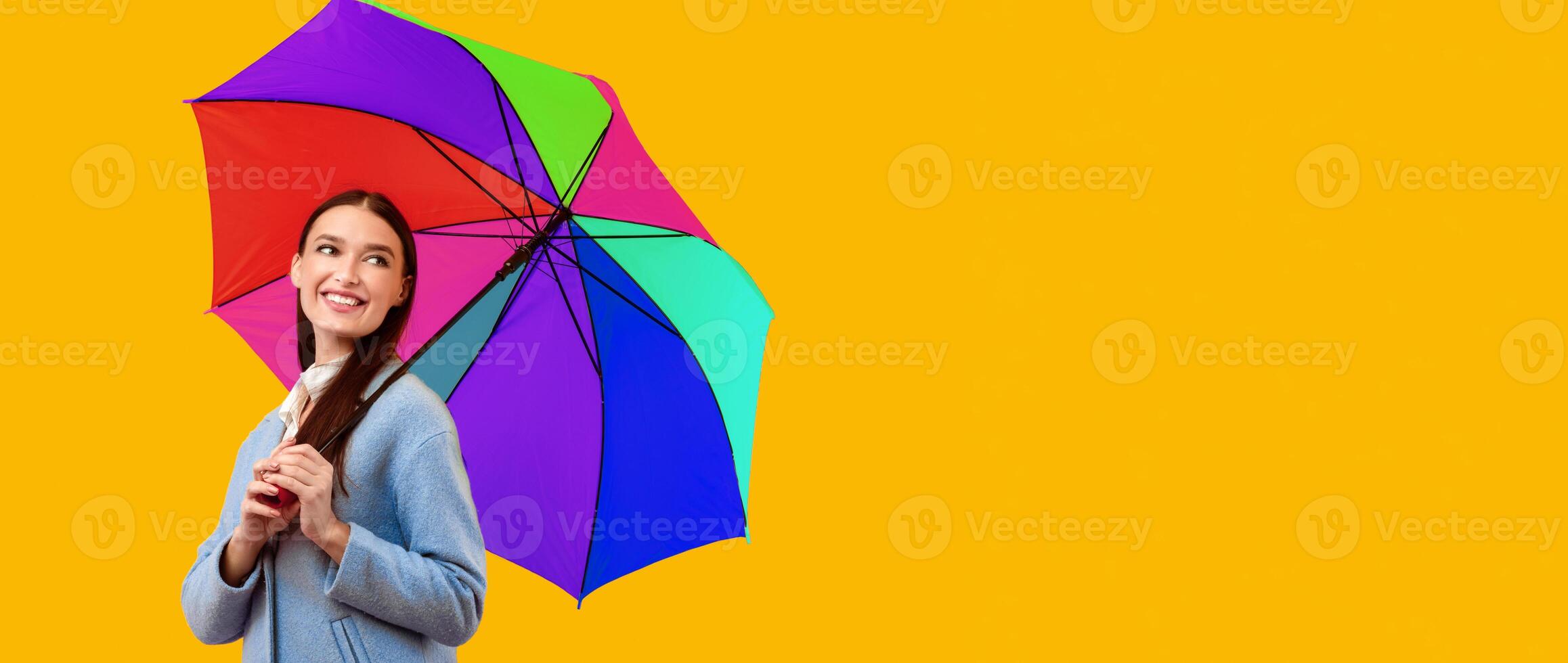 Playful Mood. Young carefree woman standing with rainbow umbrella and looking aside, yellow wall photo