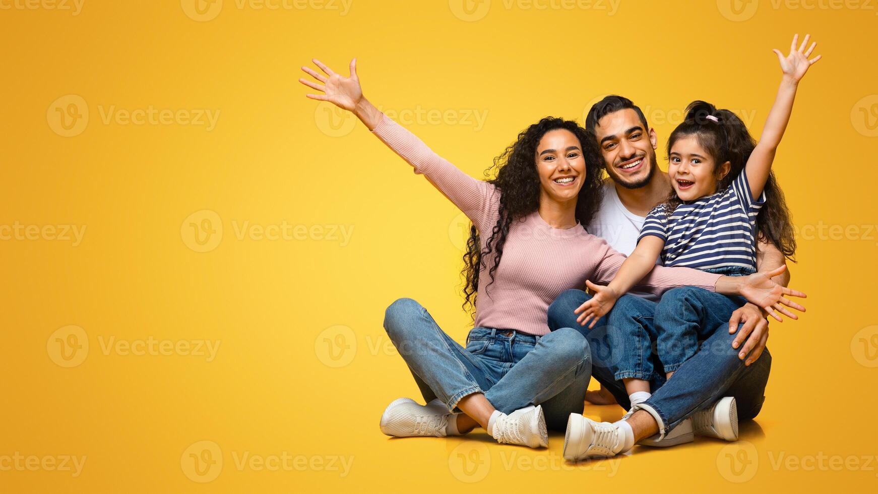 Portrait Of Happy Excited Middle Eastern Family Of Three With Little Daughter Having Fun And Bonding Together Over Yellow Studio Background, Positive Arab Parents And Child Laughing And Raising Hands photo