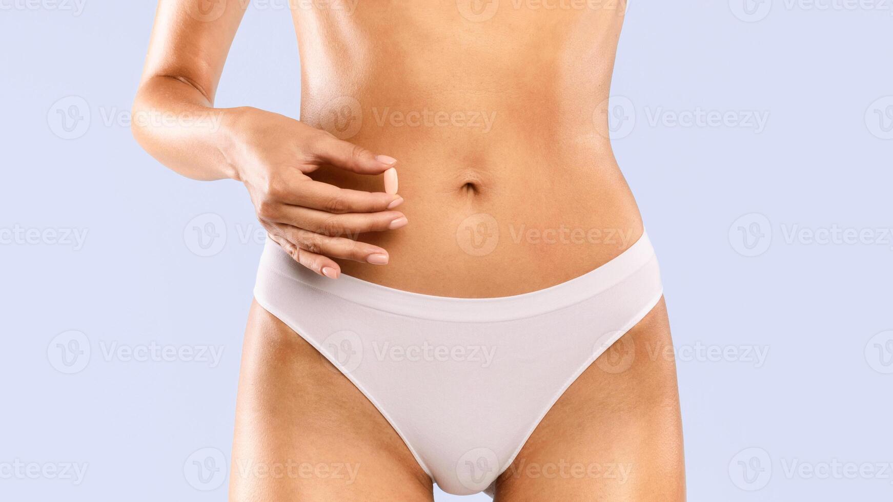 Closeup cropped view of young unrecognizable woman in white panties holding pink pill near her nude belly. Diet supplement or contraception concept, healthcare medicine treatment, studio banner photo