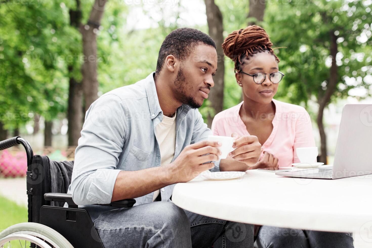 Millennial African American man in wheelchair and his female colleague using laptop together at table in outdoor cafe, having coffee while working online at park, free space photo