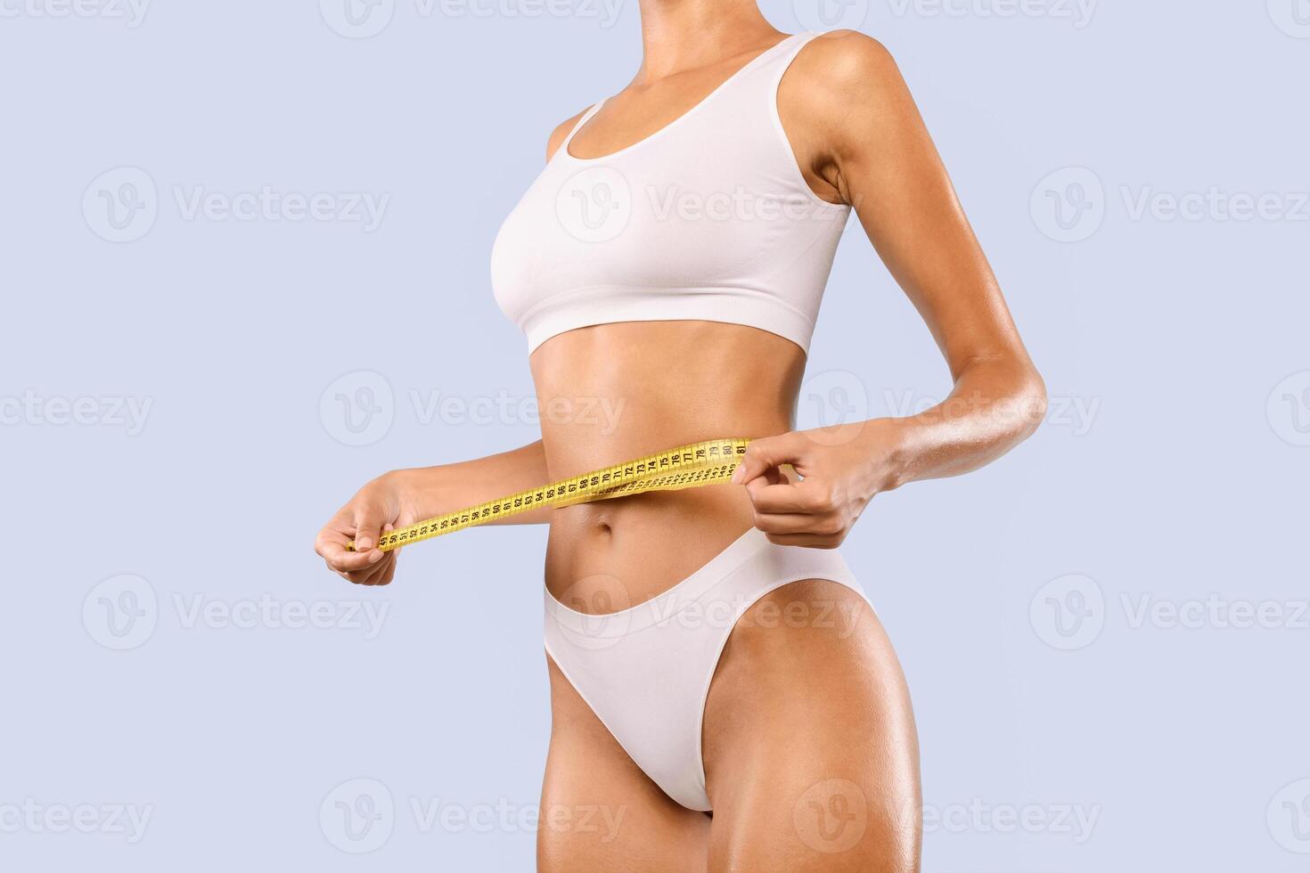 Cropped close up view of half-turned slim woman in white underwear measuring waist with yellow tape. Unrecognizable lady checking result of weightloss diet or liposuction isolated on blue studio wall photo