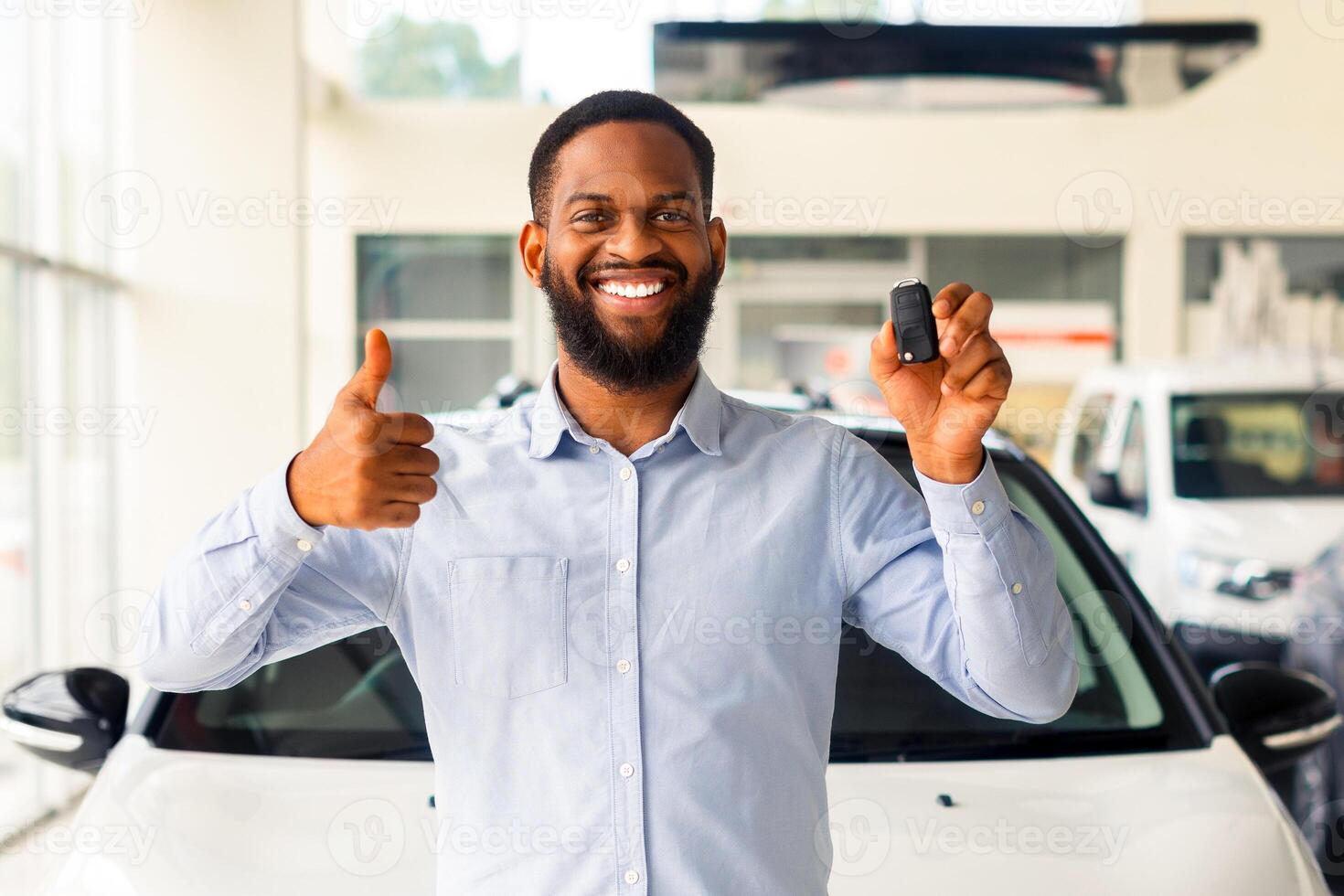 Cheerful African American Man Standing Next To His New Car, Holding Key And Showing Thumb Up. Excited Black Guy Celebrating Purchasing New Vehicle In Dealership Office, Looking And Smiling At Camera photo