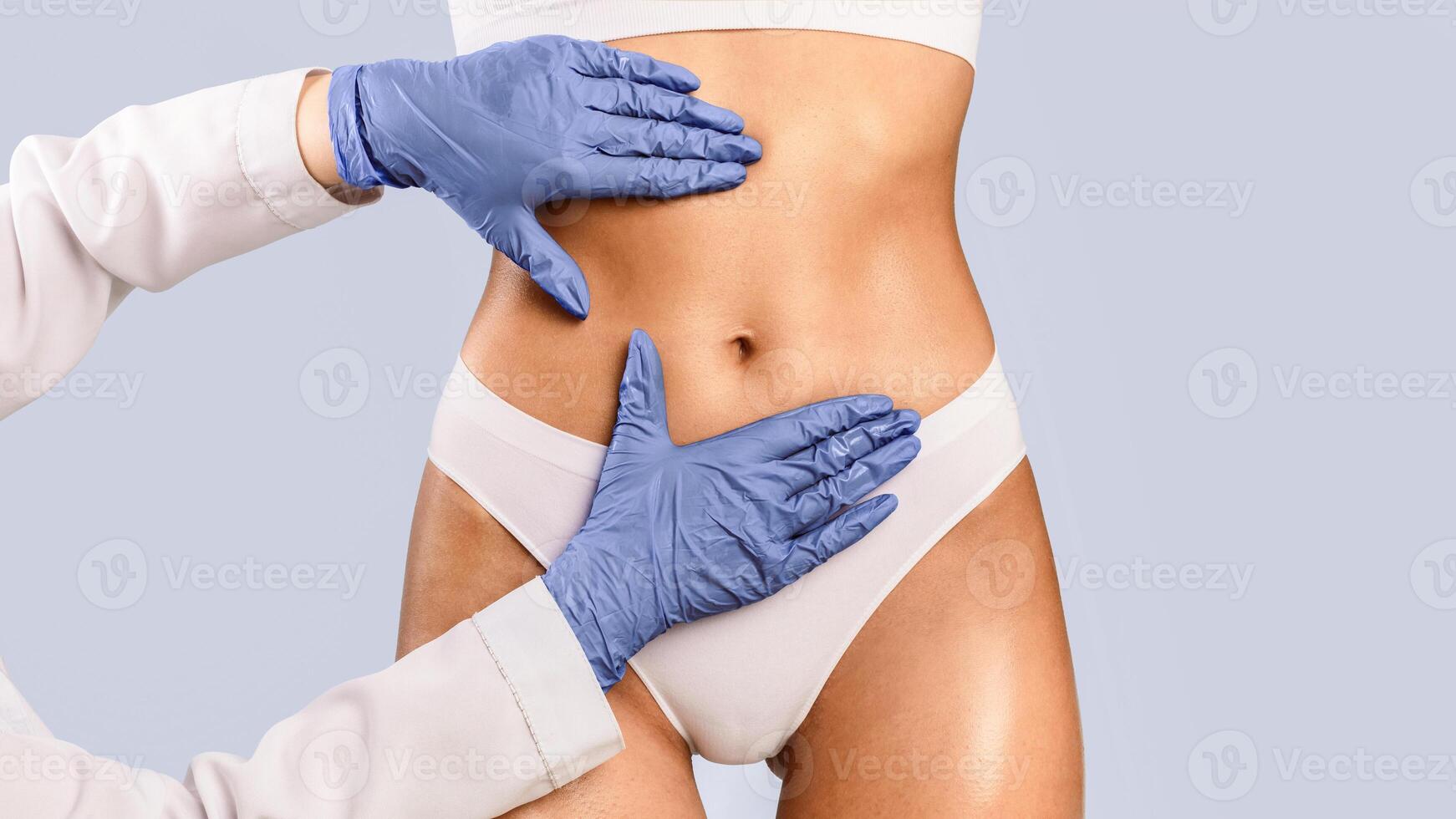 Aesthetic Medicine. Lady getting consultation at plastic surgery clinic, closeup cropped view of stomach and doctor's hands in protective gloves touching and examining belly, making fat removal plan photo