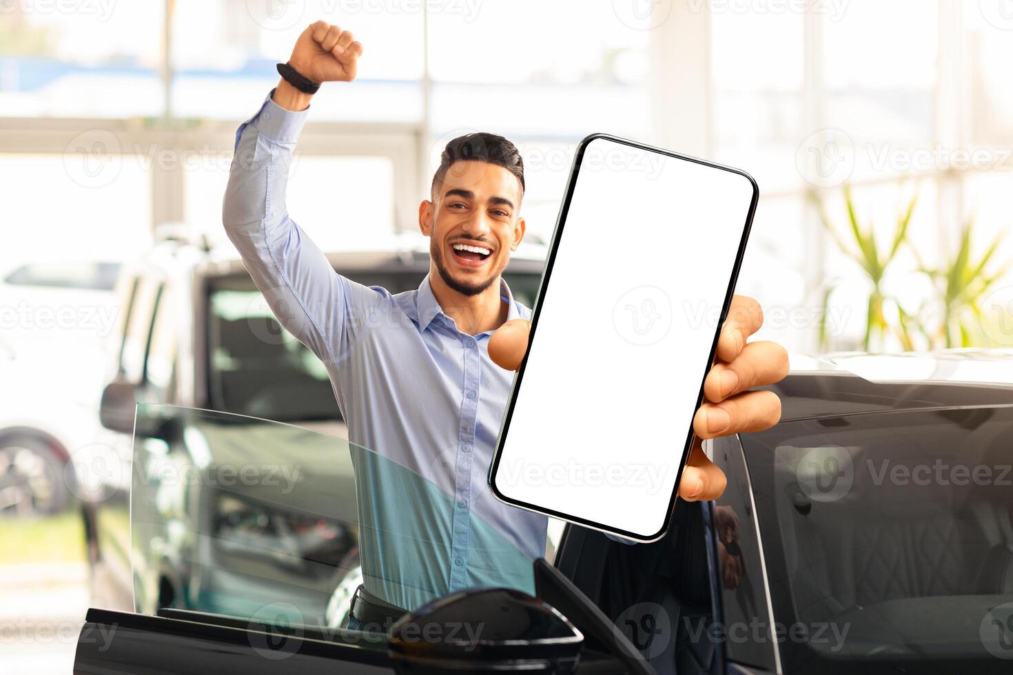 Emotional middle eastern guy choosing new car, standing by brand new black automobile, showing modern cell phone with white blank screen and raissing hand up, using auto dealership app, mockup photo