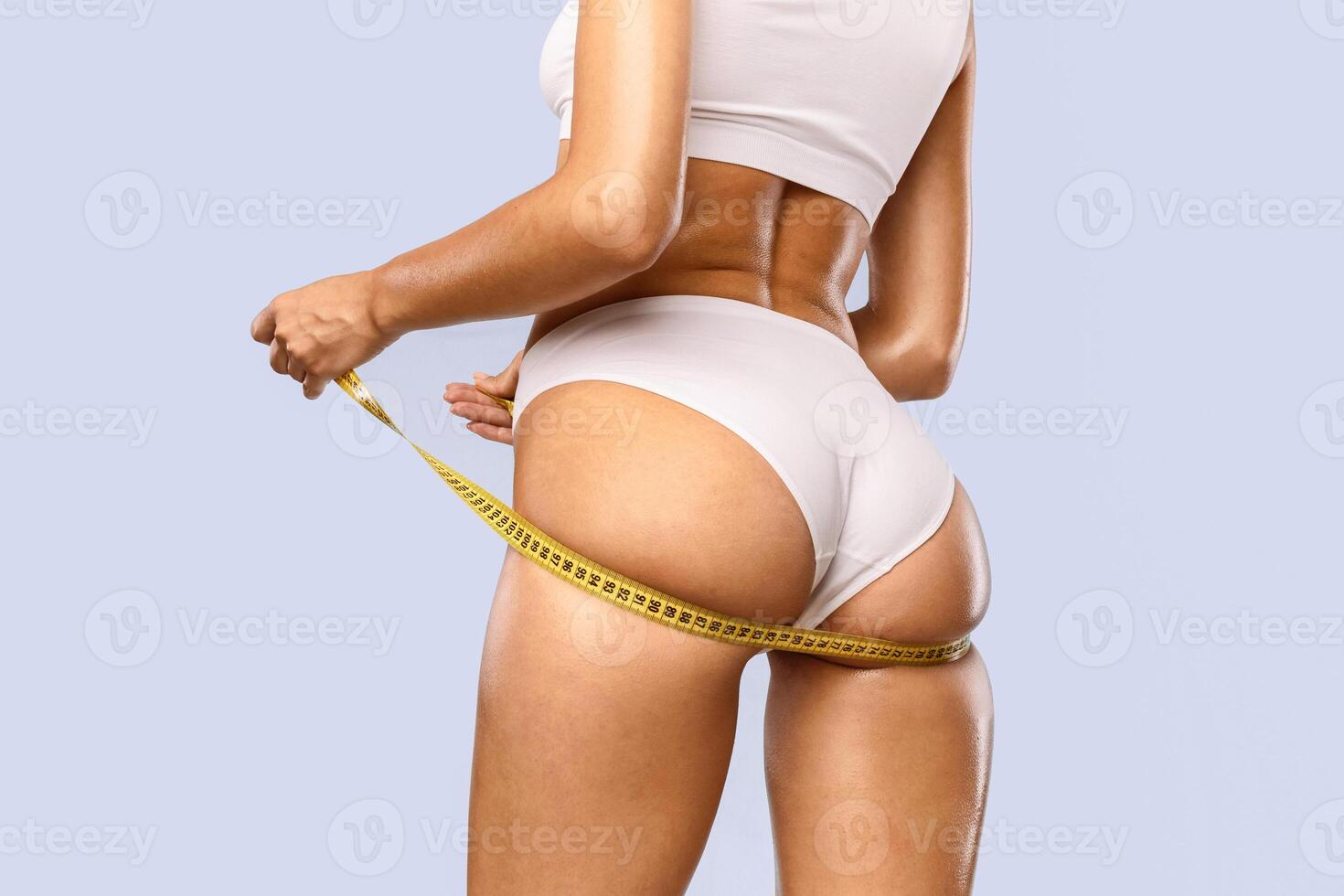 Perfect Body Concept. Cropped close up rear back view of slim woman in white bra and panties measuring her hips with yellow tape ruler. Unrecognizable lady checking her size isolated at blue studio photo